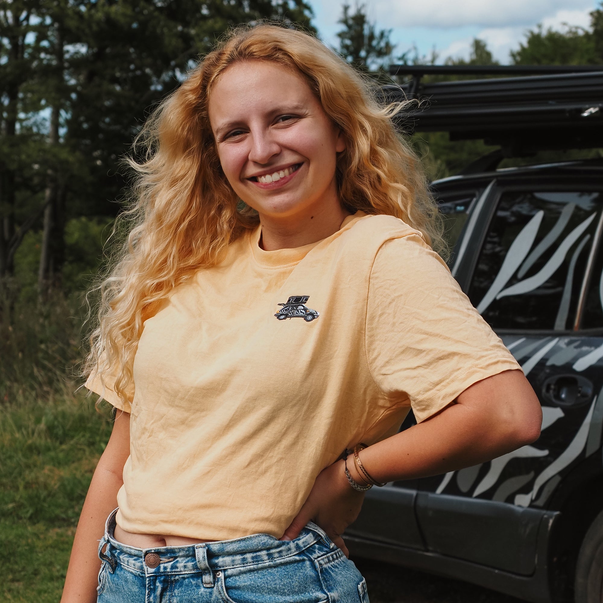 Junge Frau mit lockigem, blondem Haar trägt ein gelbes T-Shirt und lächelt vor einem gemusterten Auto.