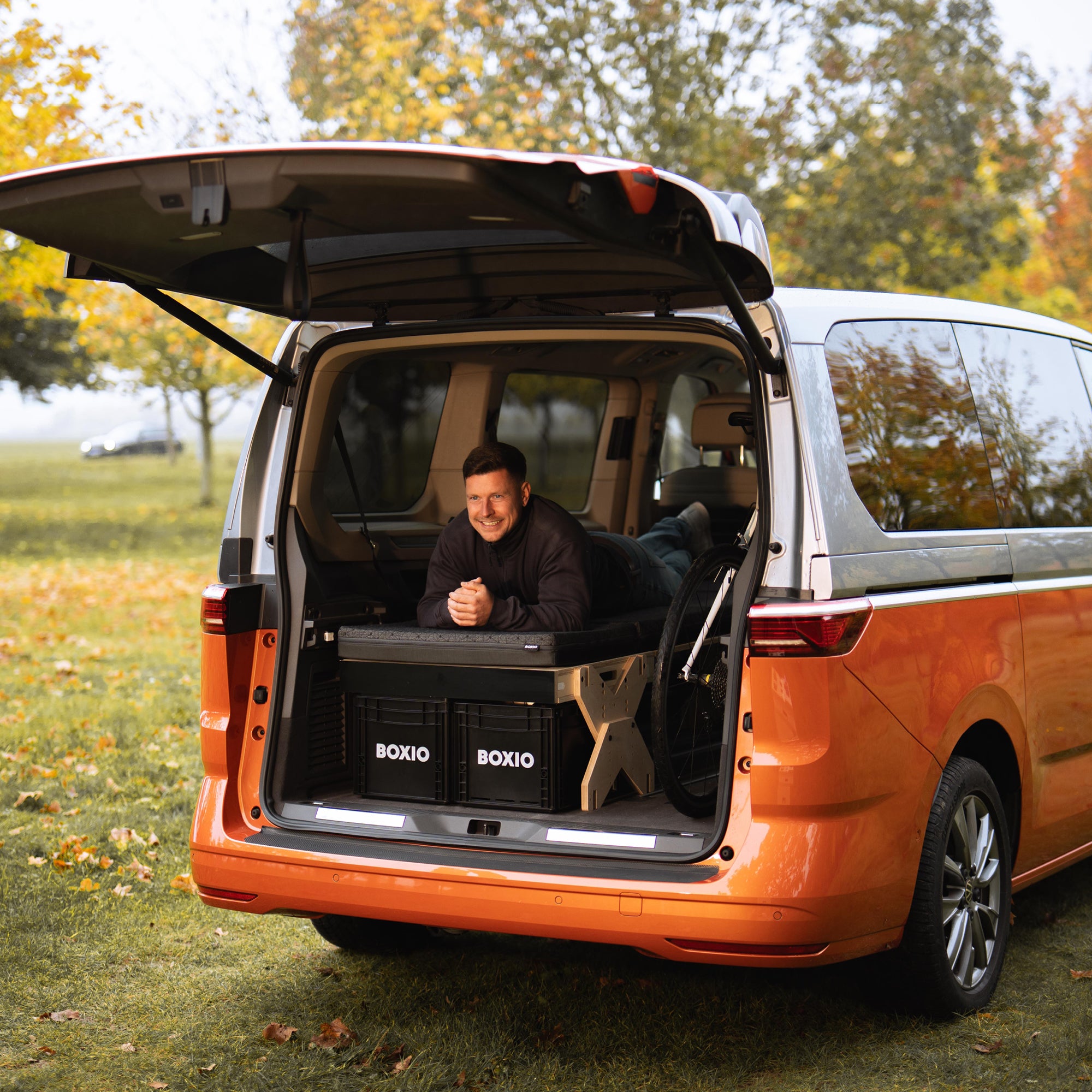 Mann liegt entspannt im Kofferraum eines orangefarbenen Vans auf einem Campingbett, umgeben von herbstlicher Natur.