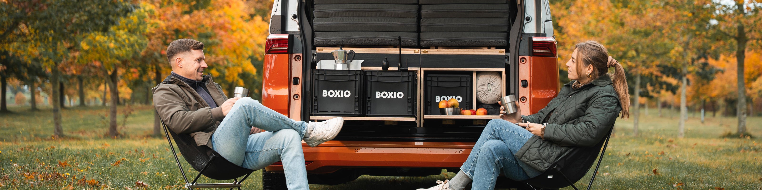 Zwei Personen sitzen entspannt vor einem Van, umgeben von buntem Herbstlaub und gemütlichen Campingutensilien.