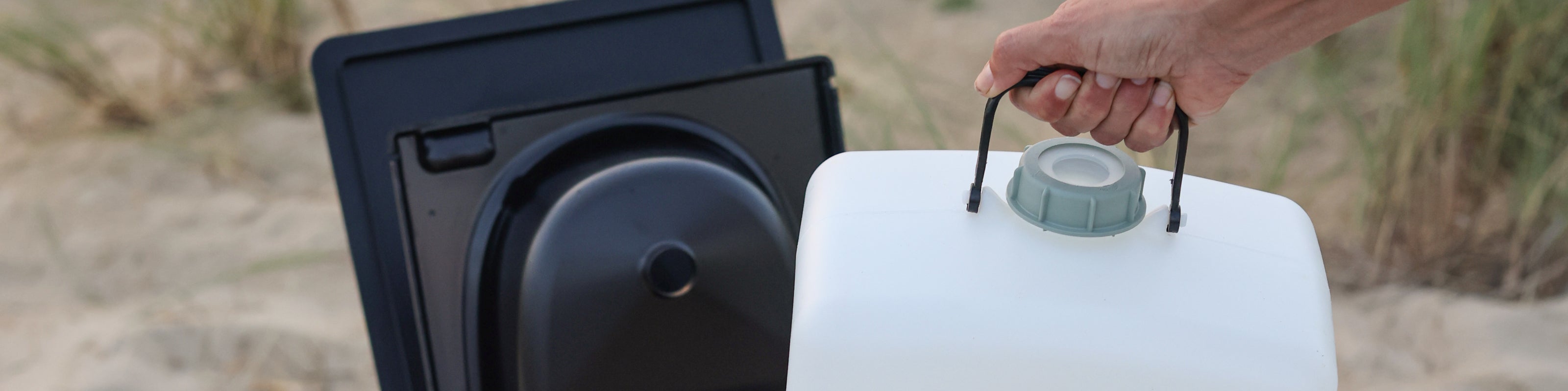 Hand hält einen weißen Wasserkanister, im Hintergrund ist ein schwarzer Behälter und Sandboden zu sehen.