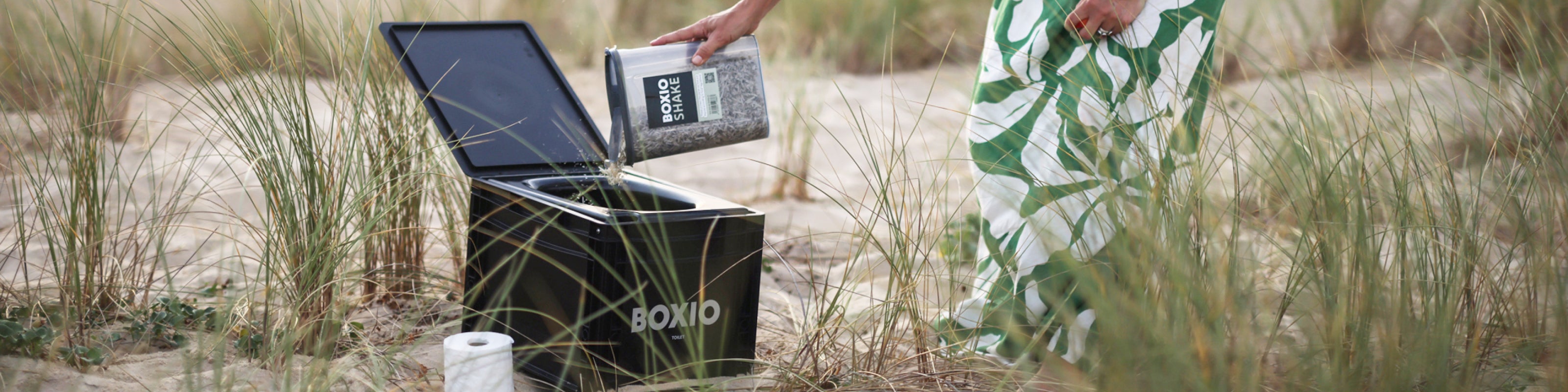 Eine Person füllt eine schwarze Boxio-Toilette mit einem grauen Produkt, umgeben von Gras und Sand.