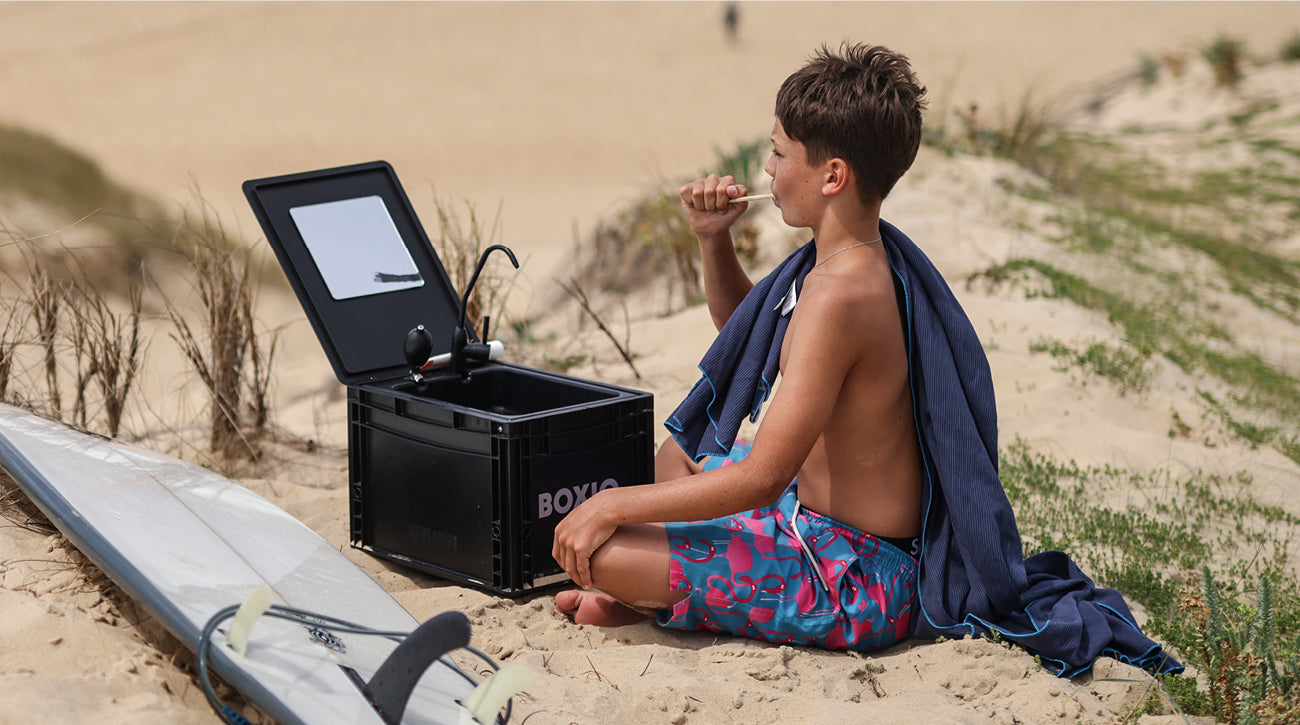 Junge sitzt am Strand, blickt auf einen offenen Kasten mit Spiegel und hält einen Snack in der Hand, neben einem Surfbrett.